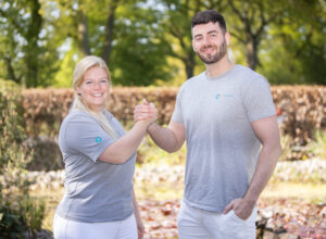 Zwei Pflegefreunde geben sich einen Handschlag und schauen dabei lächelnd in die Kamera