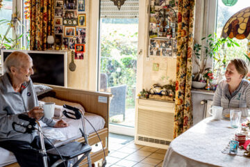 Pflegefachkraft Maria sitzt dem Patienten im Wohnzimmer gegenüber und hält eine Tasse in der Hand