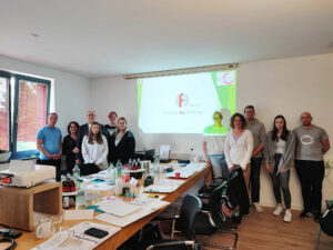 Die 9 Teilnehmer sowie Herr und Frau vom Kolke stehen aufgereiht an der Wand und schauen in die Kamera. Auf der Leinwand sieht man das Logo des Instituts für Bildung