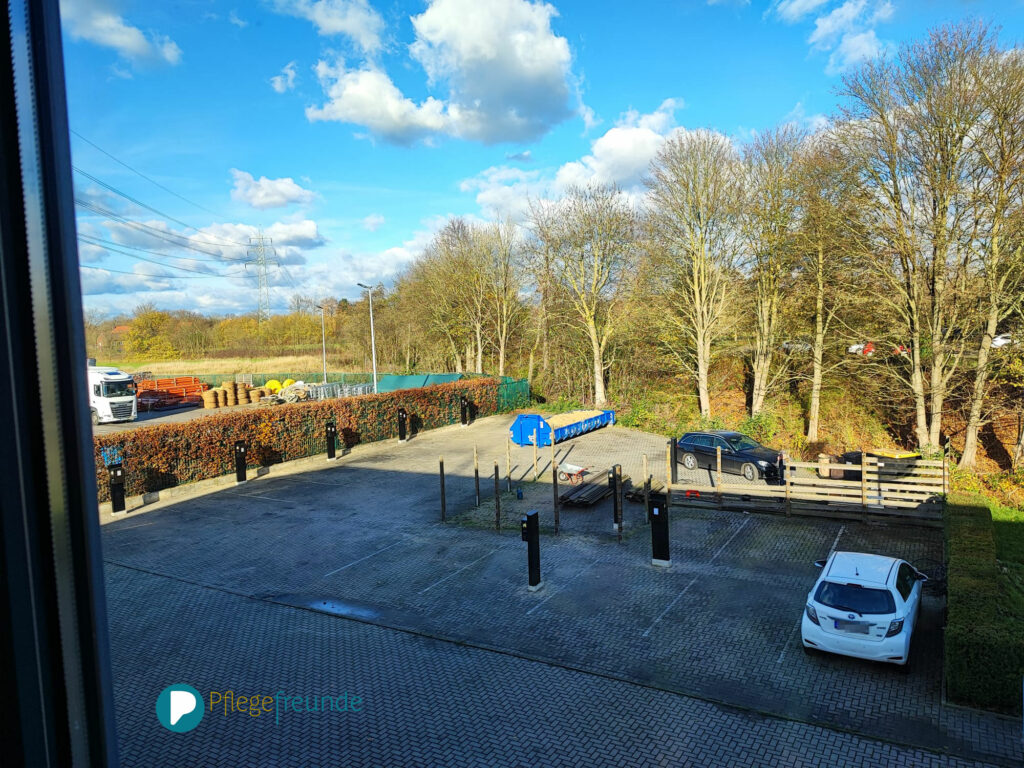 Parkplatz von oben mit Ladesäulen. Der Zaun ist halb abgerissen. Ein weißes und ein schwarzes Auto steht auf dem Parkplatz.