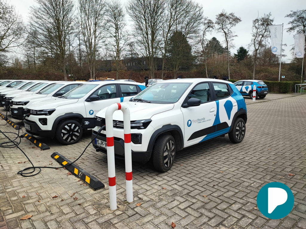 Parkplatz voll mit den E-Autos. Im Vordergrund sind zwei Autos mit blauer Folierung im Pflegefreunde-Design zu sehen.