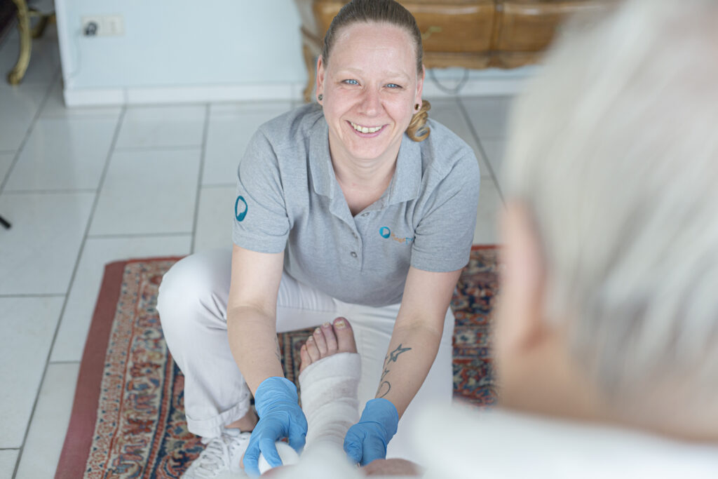 Pflegekraft versorgt das Bein eines Patienten mit einem Verband und schaut dabei in die Kamera. Patient ist von hinten zu sehen