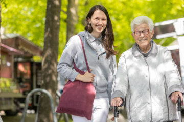 Pflegekraft und Patientin laufen gemeinsam auf die Kamera zu und lächeln hinein.