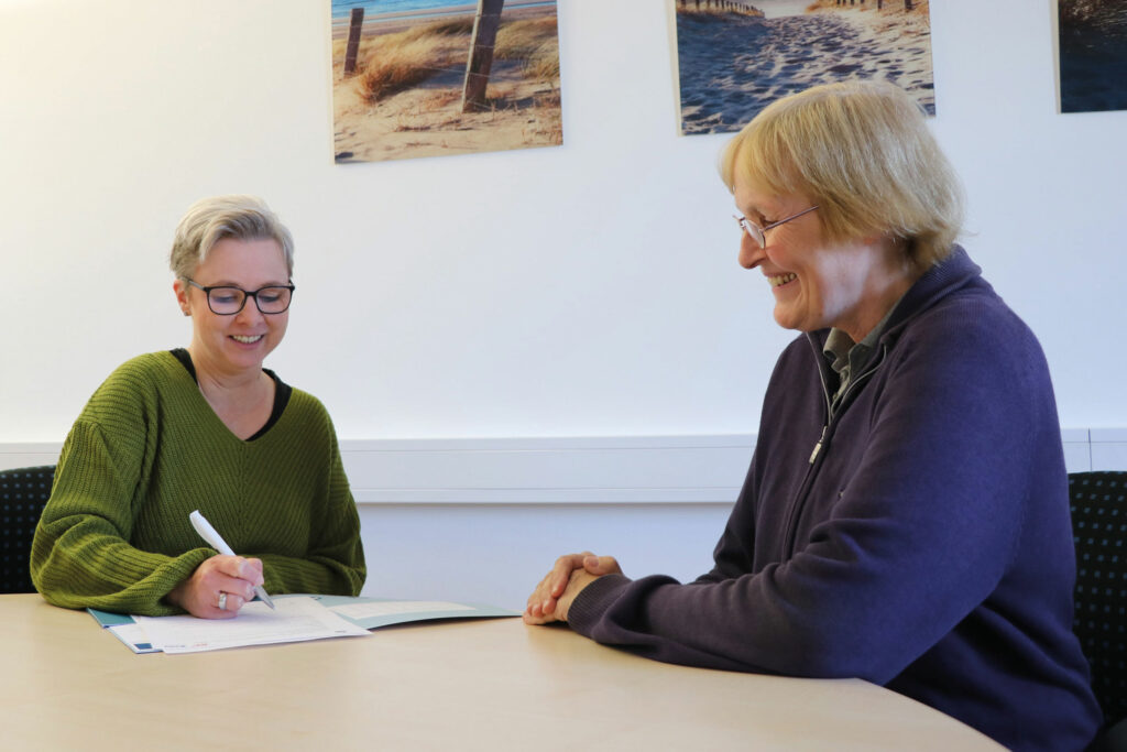 Patient und Pflegeberaterin sitzen sich lächelnd gegenüber. Pflegeberaterin schreibt etwas auf