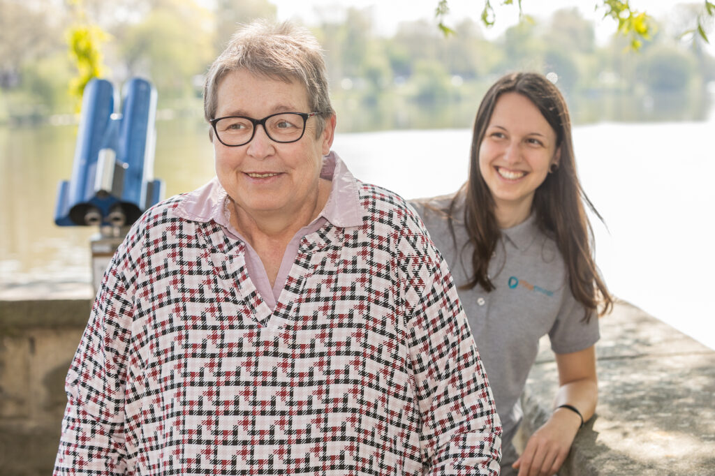 Pflegekraft steht hinter der Patientin. Beide lächeln in die Kamera