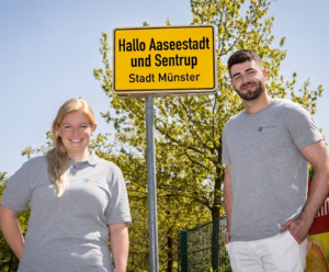 Eine Frau und ein Mann mit Pflegefreunde-Tshirt stehen vor einem gelben Ortsschild. Auf dem Schild steht "Hallo Aaseestadt und Sentrup - Stadt Münster"