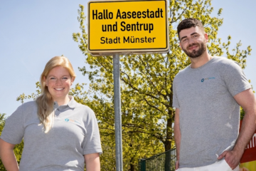 Eine Frau und ein Mann mit Pflegefreunde-Tshirt stehen vor einem gelben Ortsschild. Auf dem Schild steht "Hallo Aaseestadt und Sentrup - Stadt Münster"