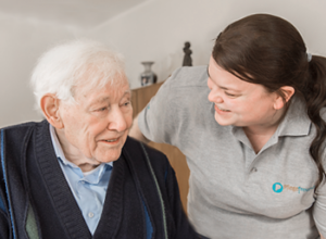 Zwei Personen auf dem Bild. Links sitzt ein älterer Herr und rechts wendet sich eine Pflegefachfrau dem Patienten zu