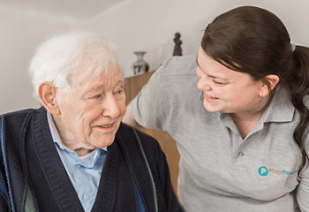 Zwei Personen auf dem Bild. Links sitzt ein älterer Herr und rechts wendet sich eine Pflegefachfrau dem Patienten zu
