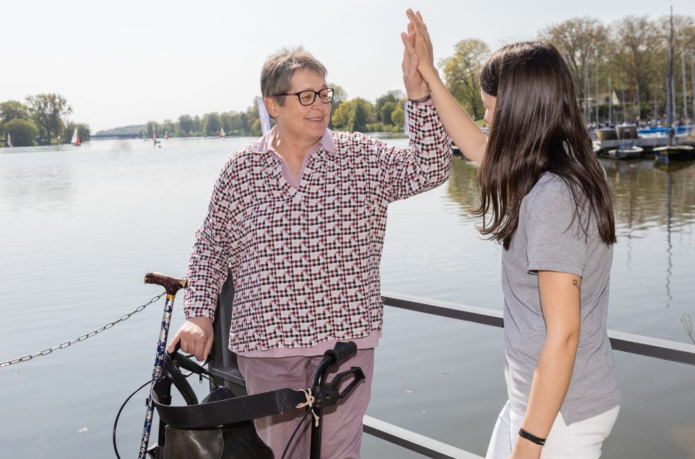 Patientin am Rollator und Pflegekraft geben sich High-Five vor dem Aasee