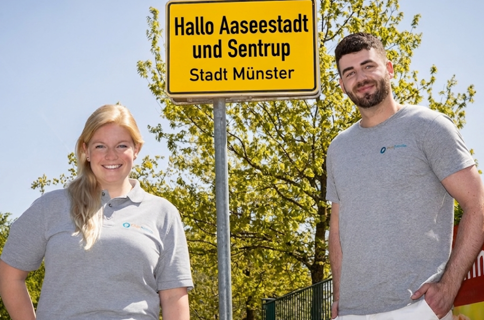 Eine Frau und ein Mann mit Pflegefreunde-Tshirt stehen vor einem gelben Ortsschild. Auf dem Schild steht "Hallo Aaseestadt und Sentrup - Stadt Münster"