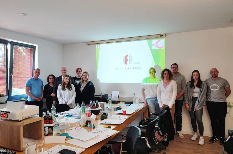 Die 9 Teilnehmer sowie Herr und Frau vom Kolke stehen aufgereiht an der Wand und schauen in die Kamera. Auf der Leinwand sieht man das Logo des Instituts für Bildung