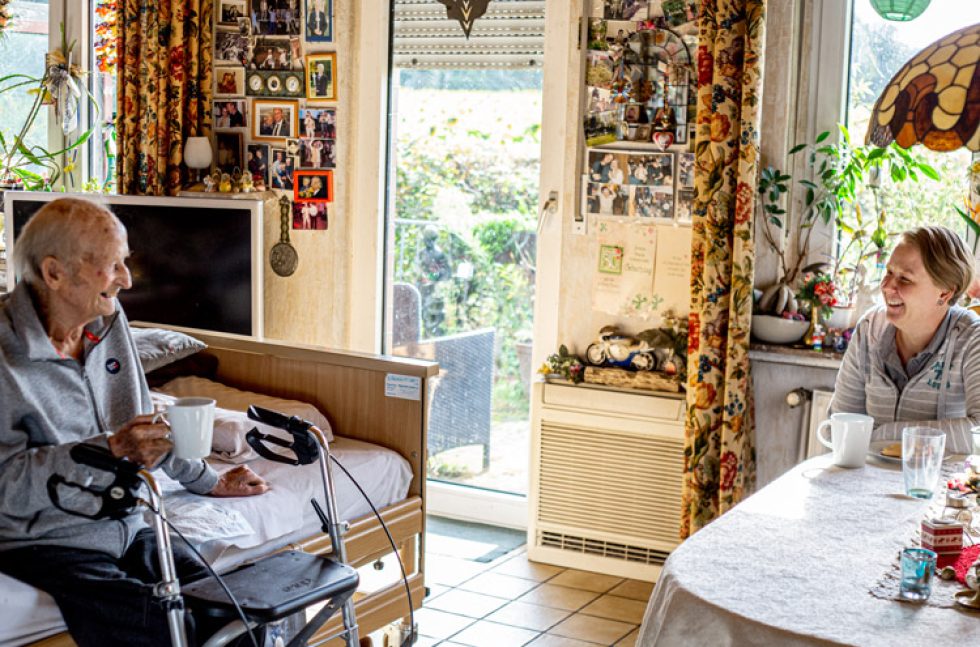 Pflegefachkraft Maria sitzt dem Patienten im Wohnzimmer gegenüber und hält eine Tasse in der Hand