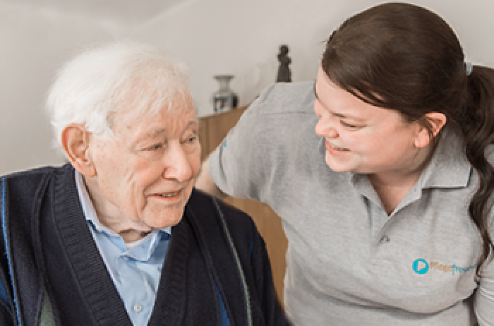 Zwei Personen auf dem Bild. Links sitzt ein älterer Herr und rechts wendet sich eine Pflegefachfrau dem Patienten zu