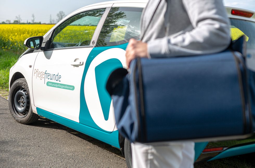 Pflegekraft mit blauer Wundtasche steht vor dem Pflegefreunde-Auto